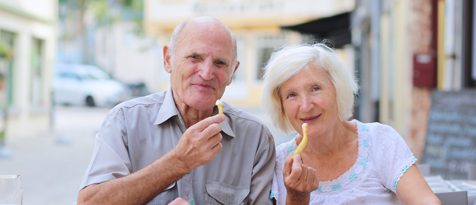 A male and female senior couple seated with each of them holding up a french fry.