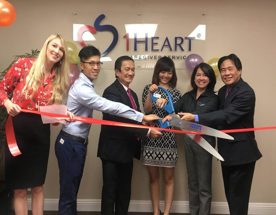 Six members of the 1 Heart Caregiver Services Team cutting a red ribbon in front of the office name sign.