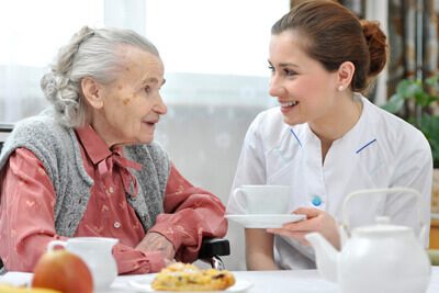A senior at the breakfast table smiling and sharing coffee with a female caregiver.