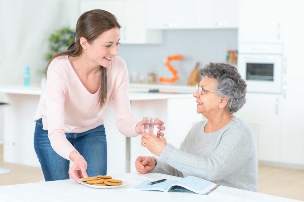 A senior seated at a kitchen table being served cookies and water by a female caregiver.
