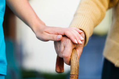 Close-up image of a senior's hand holding a cane and his hand being touched by a female caregiver's hand.