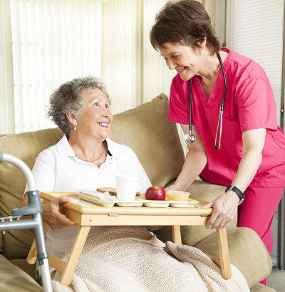 A senior woman seated on the couch being served lunch on a lap tray by her female caregiver.