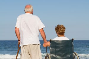 A senior standing next to his wife in a wheelchair while overlooking the beach and ocean.
