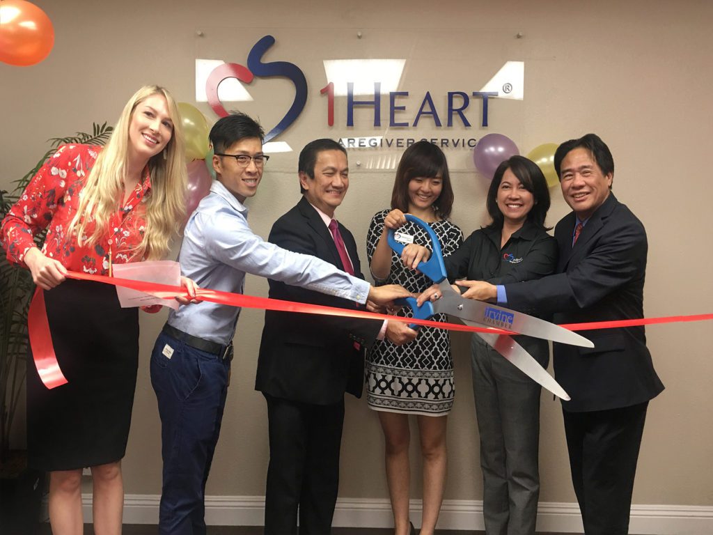 Six members of the 1 Heart Caregiver Services Team cutting a red ribbon in front of the office name sign.