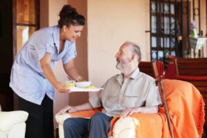A senior seated in a chair being served a meal by his female caregiver.
