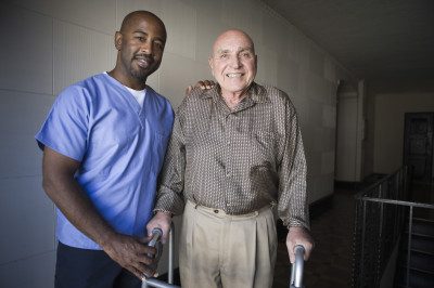 Elderly man standing with a walker being assisted by a male caregiver.