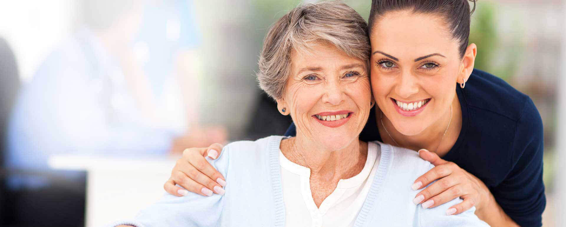 Senior woman sitting smiling next to her smiling female caregiver.