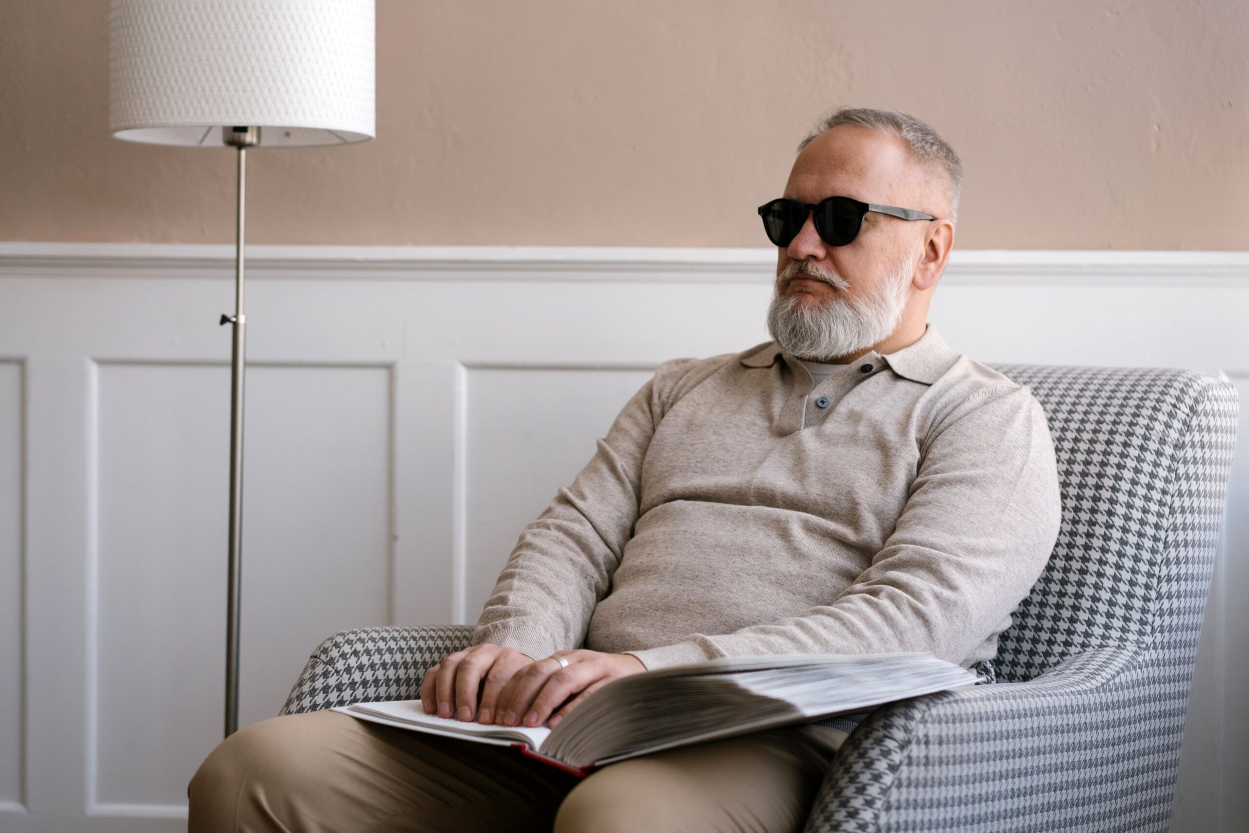 A blind elderly man sitting in an armchair, using a Braille book
