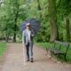 A blind elderly man with an umbrella and a walking stick, walking in a park
