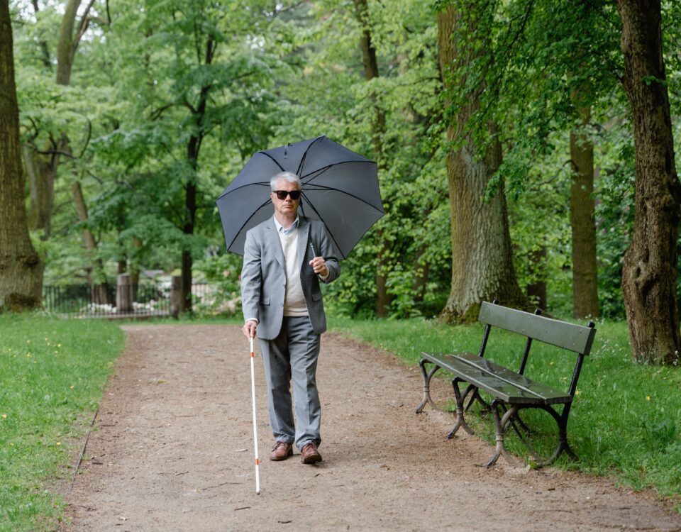 A blind elderly man with an umbrella and a walking stick, walking in a park