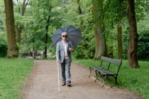 A blind elderly man with an umbrella and a walking stick, walking in a park