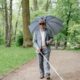 An elderly blind man walks with a cane and umbrella