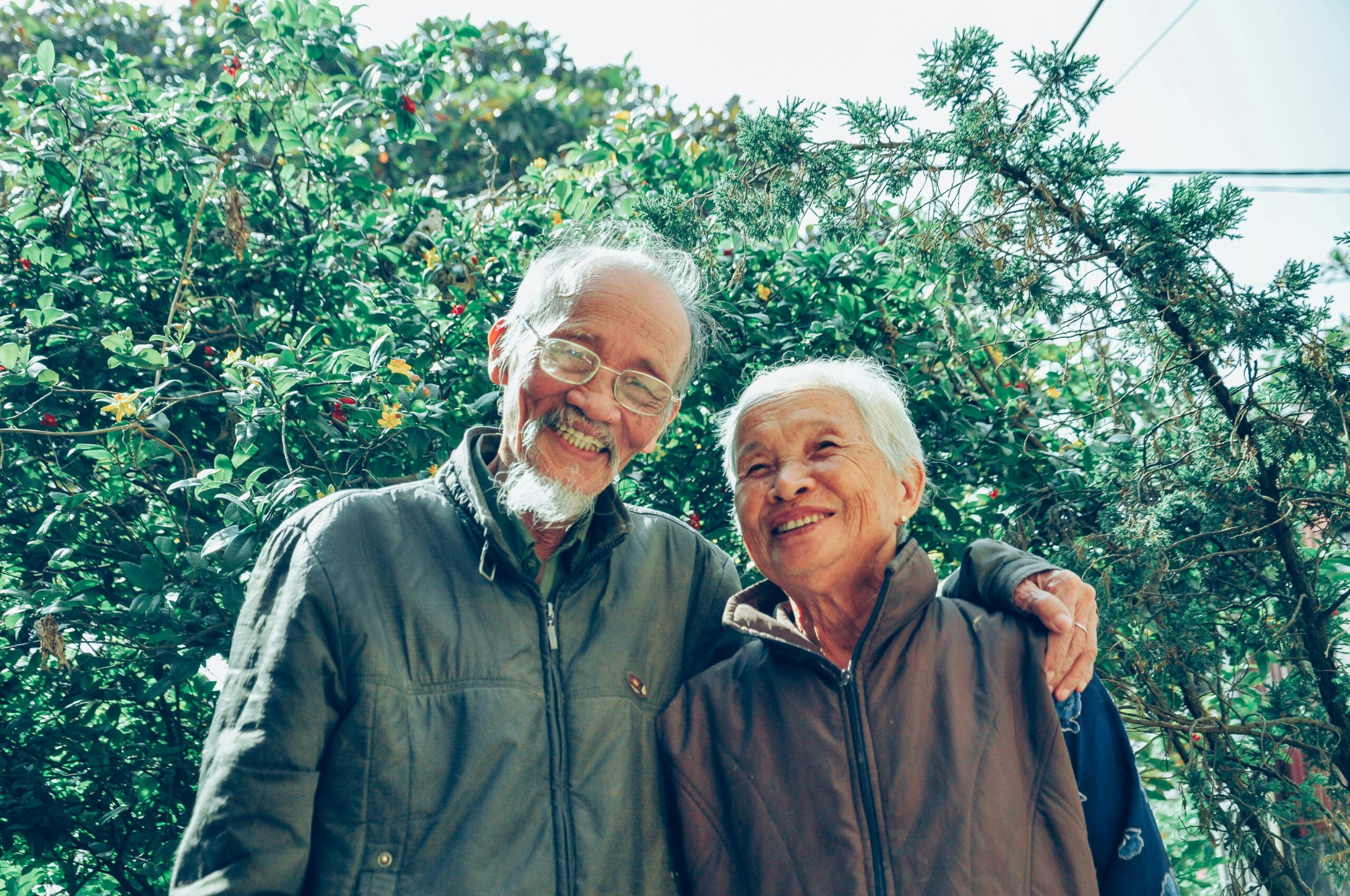 An elderly couple stands in front of a tree, smiling for the camera.